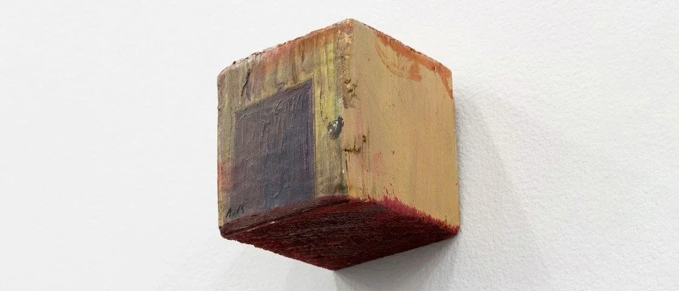 A small, weathered wooden block mounted on a white wall.